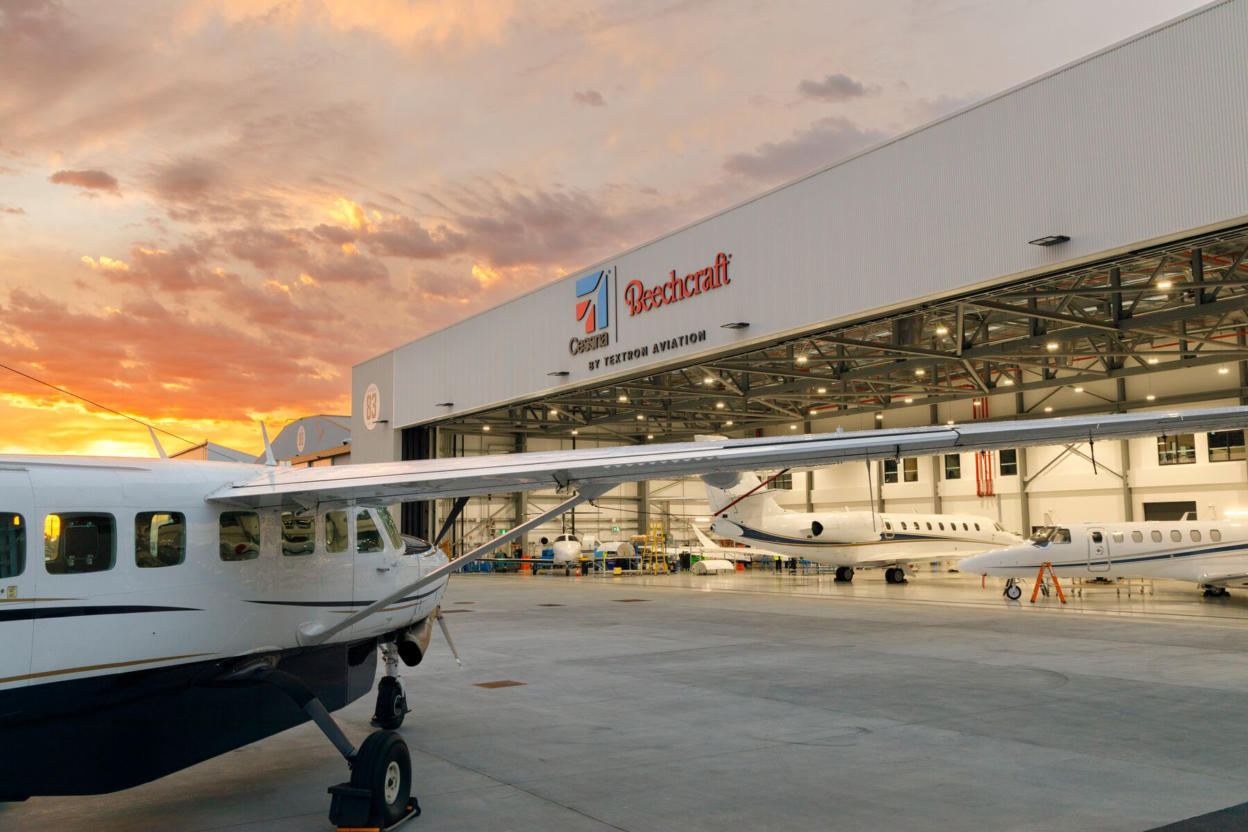 Cessna and Beechcraft aircraft parked inside and outside a large Textron Aviation hangar, with the building labeled “Cessna | Beechcraft by Textron Aviation,” under a colorful sunset sky.
